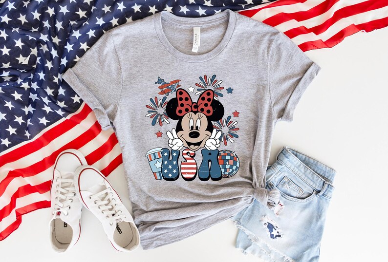 May include: A light gray t-shirt featuring a Minnie Mouse graphic with a red bow, peace signs, and the word "USA" in patriotic colors. The shirt is paired with denim shorts and white sneakers, with an American flag in the background.