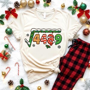 May include: Cream-colored t-shirt with a square root design featuring the numbers 4489 in festive colors. The shirt is paired with red and black plaid pajama pants, surrounded by Christmas decorations.