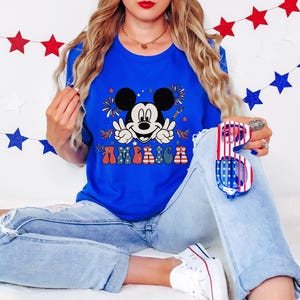 May include: A royal blue t-shirt featuring a graphic of Mickey Mouse giving the peace sign with the word "AMERICA" in colorful letters. The shirt is paired with light-wash denim jeans and patriotic sunglasses.