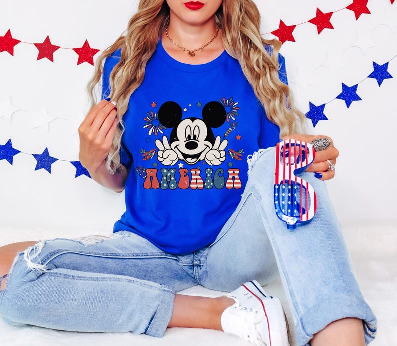 May include: A royal blue t-shirt featuring a graphic of Mickey Mouse giving the peace sign with the word "AMERICA" in colorful letters. The shirt is paired with light-wash denim jeans and patriotic sunglasses.