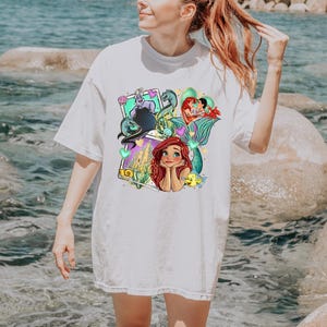A woman wearing a white t-shirt with a colorful graphic of the mermaid characters from the Disney film "The Little Mermaid".