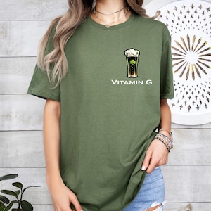 A woman wearing a green t-shirt with a graphic of a beer glass and the text "Vitamin G" printed on it, standing in front of a wooden wall with a circular design.