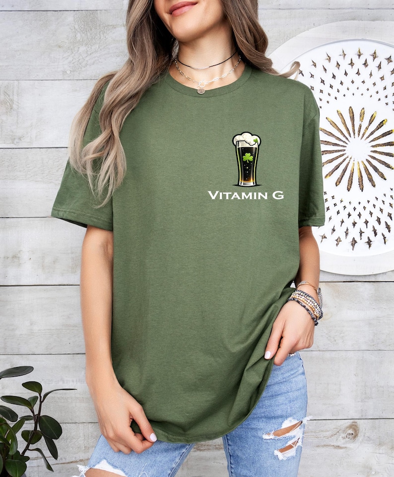 A woman wearing a green t-shirt with a graphic of a beer glass and the text "Vitamin G" printed on it, standing in front of a wooden wall with a circular design.