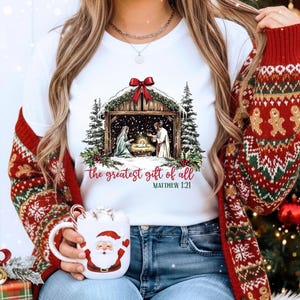 May include: White t-shirt featuring a Nativity scene with the text "the greatest gift of all" and Matthew 1:21. The image includes a red bow, evergreen trees, and a cozy Christmas theme. A Santa mug is also visible.