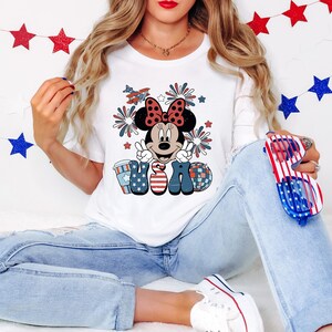 May include: White t-shirt featuring a Minnie Mouse graphic with a red bow, peace signs, and the word "USA" in red, white, and blue. The design includes fireworks and an airplane. The shirt is paired with blue jeans.