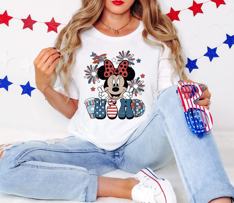 May include: White t-shirt featuring a Minnie Mouse graphic with a red bow, peace signs, and the word "USA" in red, white, and blue. The design includes fireworks and an airplane. The shirt is paired with blue jeans.