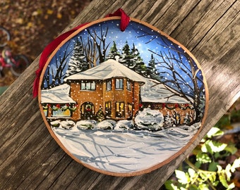May include: A hand-painted Christmas ornament depicting a brick house with a white roof and snow-covered yard. The house has a wreath on the door and lights in the windows. The ornament is surrounded by a red ribbon.