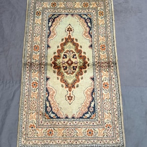 Peut inclure: Un tapis rectangulaire orné d'un médaillon central dans des tons rouille, bleu marine et crème. Le tapis présente un champ crème clair avec des motifs floraux complexes, une bordure multicouche à motifs de fleurs orange et des franges blanches.