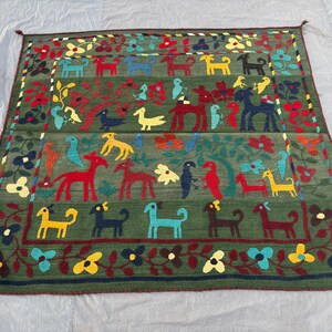 May include: A rectangular, hand-woven rug with a green background and a border of flowers and vines. The rug features rows of colorful animal and bird figures, including dogs, horses, and birds. The design incorporates red, yellow, blue, and black elements.