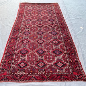 Peut inclure: Un tapis rectangulaire tissé à la main, avec un fond rouge profond et des motifs géométriques complexes. Le design présente des formes de losanges et hexagonales en rouge, violet et noir, avec une bordure détaillée. Le tapis est probablement en laine.