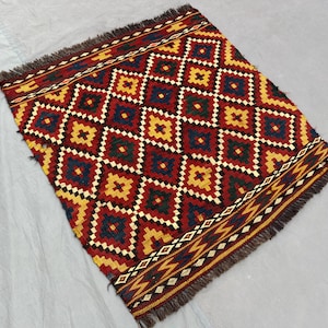 Puede incluir: Una alfombra rectangular tejida a mano con un patrón geométrico de diamantes. La alfombra presenta un patrón repetitivo de diamantes rojos, amarillos, azules y verdes sobre un fondo rojo. Los bordes están acabados con flecos.