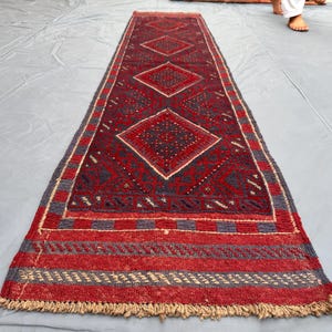 Peut inclure: Un tapis rectangulaire tissé à la main avec un motif de losanges répété dans des tons de rouge, bleu marine et beige. Le tapis présente une bordure à franges et est probablement en laine. Le design est symétrique et traditionnel.