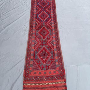 Peut inclure: Un tapis couloir rectangulaire rouge et gris avec un motif géométrique en losanges. Le tapis présente un motif central de losanges rouges et bleus répétés, encadrés de bordures à motifs complexes et de franges.