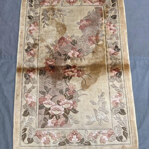 Peut inclure: Un tapis rectangulaire au fond beige orné d'un motif floral central dans des tons de rose, brun et crème. Le tapis présente une bordure décorative avec des motifs de fleurs répétitifs et des franges sur les bords supérieur et inférieur.