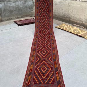 Peut inclure: Un long tapis rectangulaire avec un motif géométrique en losanges dans des tons de rouge, d'orange et de jaune. Le tapis a une frange en bas et est exposé à l'extérieur. Le tapis mesure environ 300 cm de long.