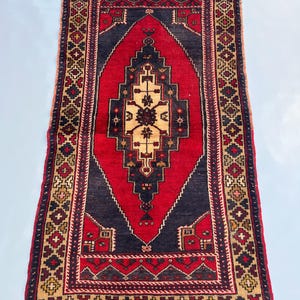 Puede incluir: Una alfombra rectangular tejida a mano con un patrón central de diamante en rojo y beige, enmarcada por un borde azul marino. La alfombra presenta intrincados diseños geométricos en tonos de rojo, beige y azul marino, con un borde con flecos.