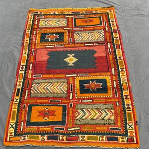 Peut inclure: Un tapis rectangulaire présentant un motif géométrique vibrant avec un panneau central bleu foncé contenant un motif en losange blanc, entouré de blocs rectangulaires alternés orange et bleu avec des motifs d'étoiles et de chevrons en rouge et jaune.