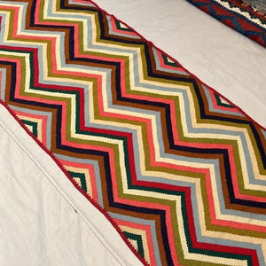 Peut inclure: Un tapis de couloir à motifs en zigzag multicolores. Le tapis présente un motif de chevrons répété dans des tons de rouge, rose, vert, bleu et marron. Les bords sont finis avec une bordure rouge. Le tapis est probablement en tissu tissé.