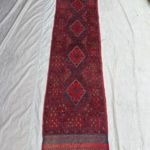 Peut inclure: Un long tapis rectangulaire rouge avec un motif géométrique. Le tapis présente un motif de losanges répétitifs dans des tons de bleu et de rouge, avec une bordure de rayures de couleurs similaires. Le tapis est probablement en tissu tissé.