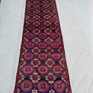 Puede incluir: Una alfombra larga y rectangular con un fondo azul oscuro y un patrón repetitivo de formas geométricas rojas y beige. La alfombra tiene flecos en ambos extremos y probablemente está hecha de lana.