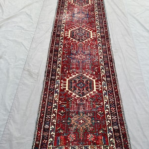Peut inclure: Un long tapis rectangulaire rouge avec des motifs géométriques complexes en bleu, blanc et vert. Le tapis présente un motif central avec des formes répétitives et une bordure détaillée. Des franges sont visibles aux extrémités.