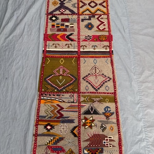 Peut inclure: Un tapis rectangulaire tissé à la main avec un fond beige et une série de motifs géométriques rouges, jaunes, verts et bleus. Le tapis a une bordure rouge et des franges aux extrémités. Le design est divisé en sections rectangulaires.