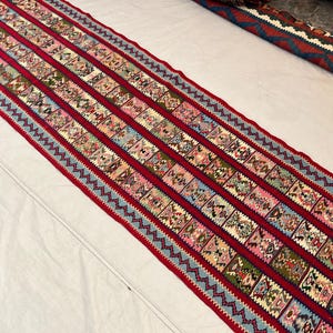 Peut inclure: Un long tapis rectangulaire au design vibrant. Le tapis présente un champ central rouge foncé avec des rangées de carrés à motifs de différentes couleurs, dont le rose, le bleu et le vert. Les bords sont bordés d'un motif en zigzag bleu clair, créant un contraste saisissant.