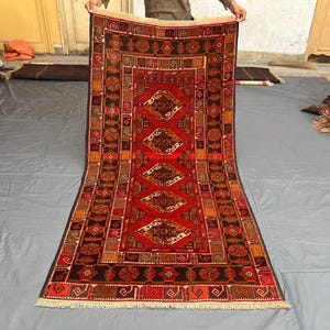 Peut inclure: Un tapis rectangulaire tissé à la main avec un champ central rouge et des motifs géométriques complexes en marron, orange et crème. Le tapis présente un motif en forme de losange et une bordure à franges.