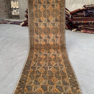 Peut inclure: Un tapis rectangulaire à motifs géométriques bruns et orangés. Le tapis a une bordure marron foncé et une frange crème. Le tapis est tenu, montrant sa longueur et son design. Le tapis est en tissu.