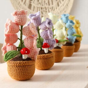 May include: Five crocheted flower pots with different colored flowers. Each pot has a small red mushroom at the base. The pots are brown and the flowers are pink, purple, yellow, light blue, and a darker blue.
