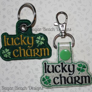 May include: Two keychains with the text "lucky charm" embroidered on them. One keychain is green with gold lettering and the other is white with black lettering. Both keychains have a shamrock design.