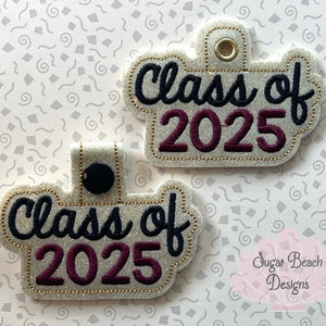 May include: Two white glitter keychains with black and burgundy embroidery. The keychains are shaped like a cloud and say "Class of 2025".