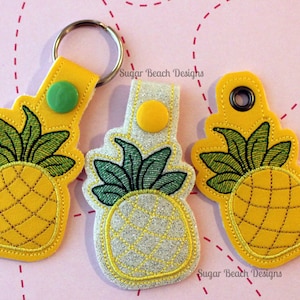 May include: Three pineapple-shaped keychains with yellow fabric and green embroidered leaves. The keychains have different embellishments: a green button, a yellow button, and a metal ring.