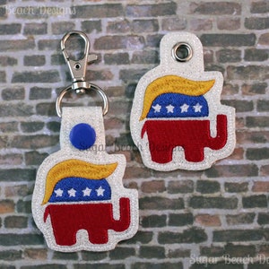 May include: Two red, white, and blue elephant keychains with a yellow hairpiece on top. The keychains are embroidered with stars and are attached to silver key rings.