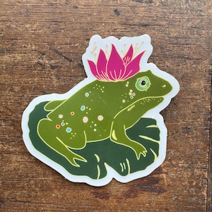 May include: A green frog sticker with a pink flower crown. The frog is sitting on a green lily pad. The sticker has a white border and is on a wooden surface.
