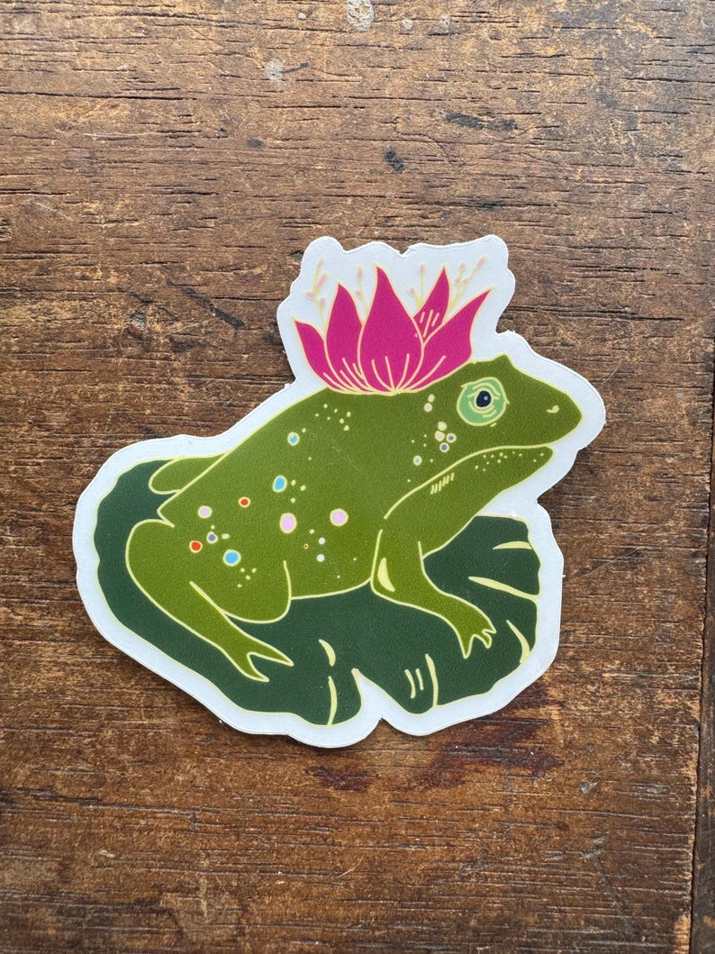 May include: A green frog sticker with a pink flower crown. The frog is sitting on a green lily pad. The sticker has a white border and is on a wooden surface.