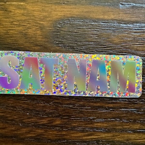 May include: A holographic sticker with the word "SAT NAM" in a rainbow color scheme.