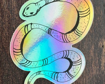 May include: A holographic sticker featuring a black and white line drawing of a snake curled up in a circle. The sticker has a rainbow iridescent finish.