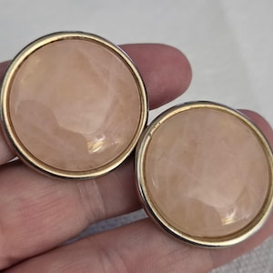 May include: A pair of round earrings featuring smooth, pale peach-colored cabochons set within polished gold-tone metal frames. The circular stones have a subtle, marbled texture and a glossy finish, creating a simple and uniform appearance.