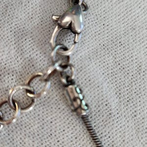 May include: Silver-toned necklace with a snake chain and decorative clasps. The necklace features a lobster clasp and a small chain for adjusting the length. The design includes decorative elements on the chain ends.