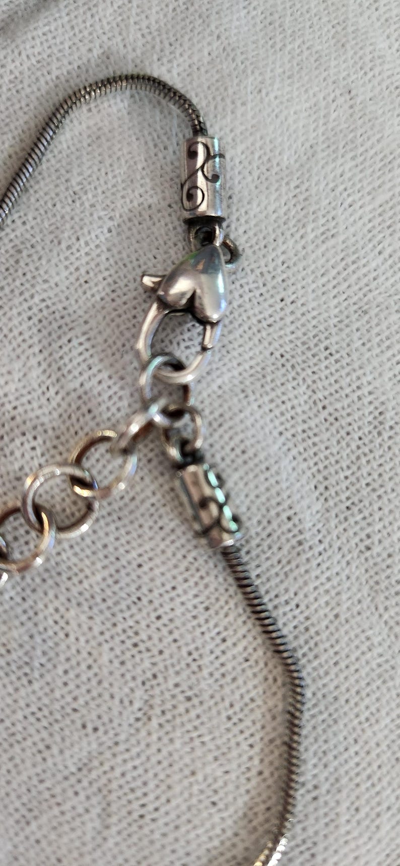 May include: Silver-toned necklace with a snake chain and decorative clasps. The necklace features a lobster clasp and a small chain for adjusting the length. The design includes decorative elements on the chain ends.