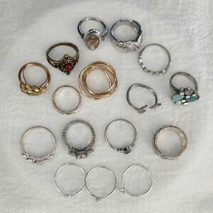 May include: A collection of various rings, including gold, silver, and gemstone designs. The rings feature different styles, such as bands, cocktail rings, and rings with intricate details. The gemstones include clear, red, and turquoise stones.