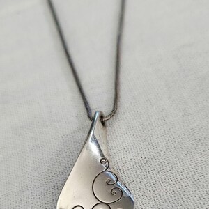 May include: Silver-toned pendant necklace with a teardrop-shaped pendant. The pendant features a swirling, scroll-like design on the lower right side. The necklace chain is a thin, dark silver color.