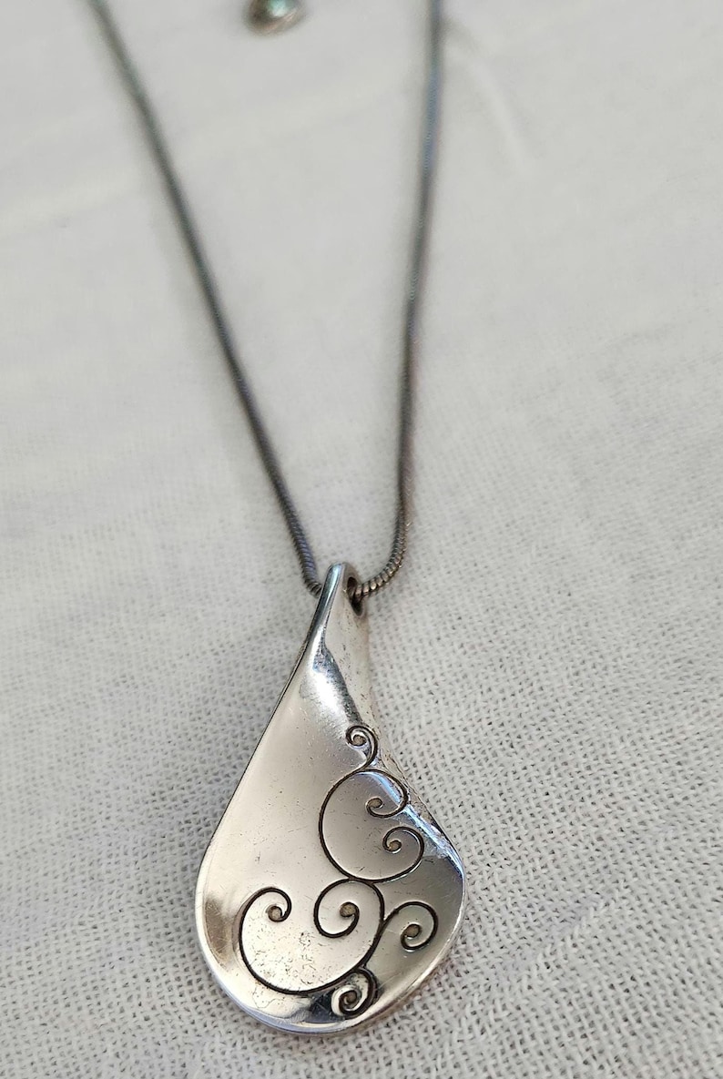 May include: Silver-toned pendant necklace with a teardrop-shaped pendant. The pendant features a swirling, scroll-like design on the lower right side. The necklace chain is a thin, dark silver color.