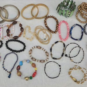May include: A collection of assorted bracelets including metal bangles, beaded strands in various colors, textures, and materials, a green rectangular link bracelet, and a gold-toned tree charm bracelet, all arranged together on a neutral surface.