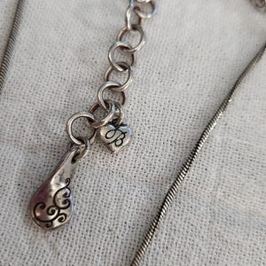 May include: Silver-toned necklace with a teardrop pendant and a heart-shaped charm. The teardrop pendant has an ornate design, and the heart charm features the letter "B". The necklace has a delicate chain.