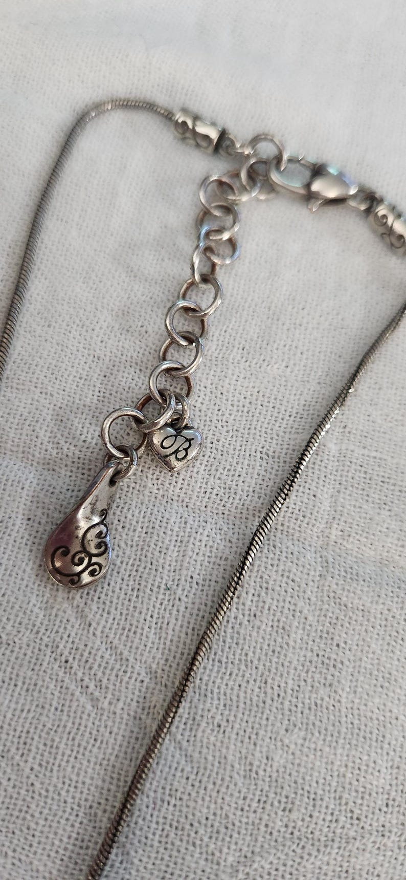 May include: Silver-toned necklace with a teardrop pendant and a heart-shaped charm. The teardrop pendant has an ornate design, and the heart charm features the letter "B". The necklace has a delicate chain.