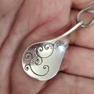 May include: A silver pendant with a teardrop shape and a decorative swirl design. The pendant is attached to a silver chain. The design is etched into the metal, creating a contrast between the shiny surface and the detailed pattern.