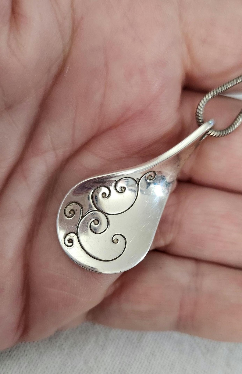 May include: A silver pendant with a teardrop shape and a decorative swirl design. The pendant is attached to a silver chain. The design is etched into the metal, creating a contrast between the shiny surface and the detailed pattern.