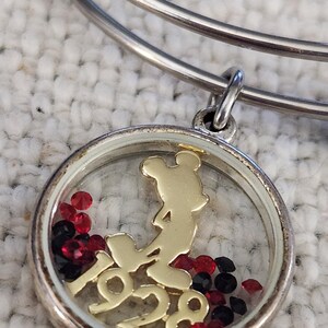 Puede incluir: Pulsera de brazalete plateada con un dije redondo. El dije presenta una silueta dorada de Mickey Mouse y el a&ntilde;o "1928". Cristales rojos y negros est&aacute;n dentro del dije. La pulsera est&aacute; sobre una superficie blanca texturizada.